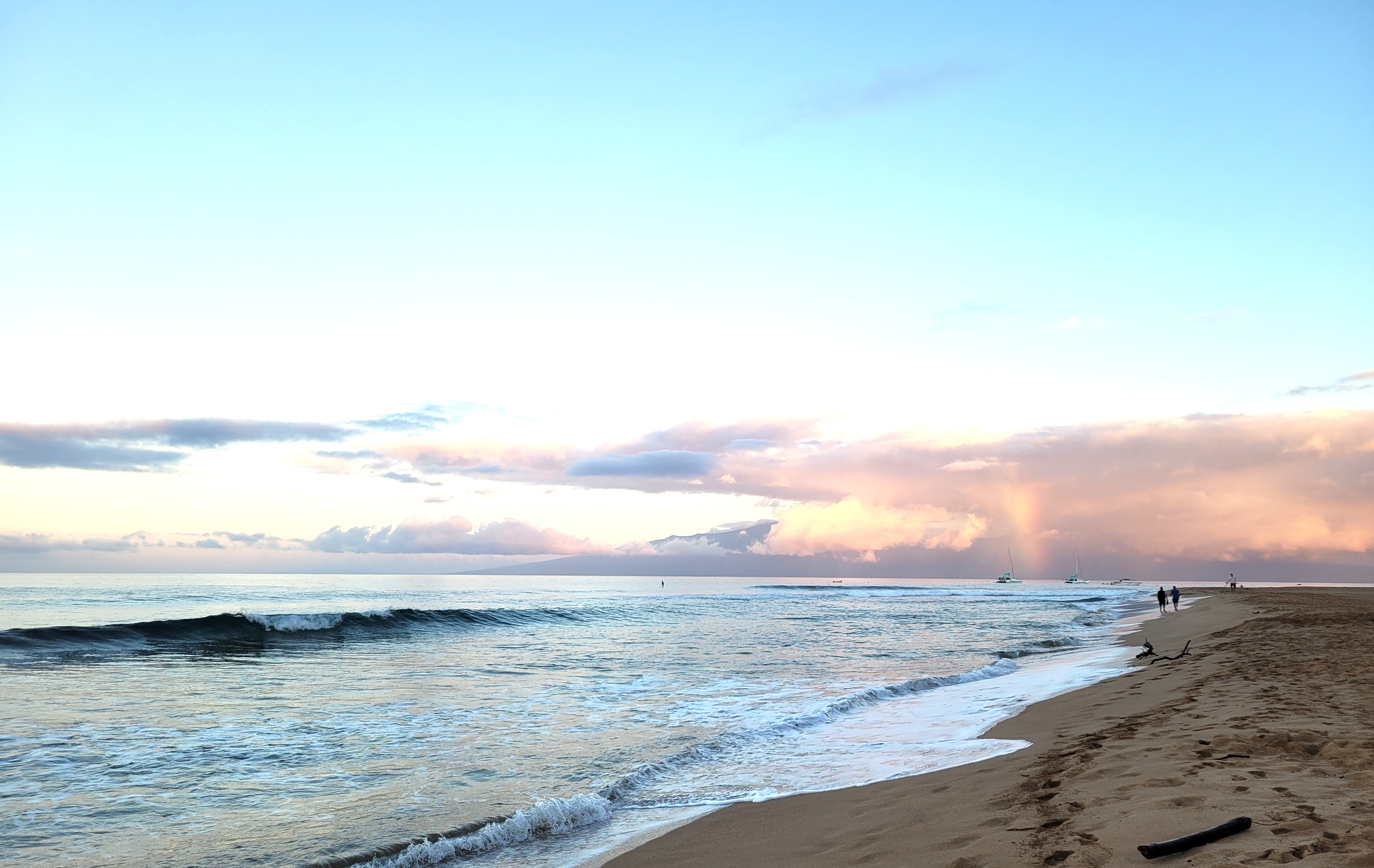 Coastal view at sunset with gentle waves crashing on the shore and pink clouds in the sky, two figures walking along the beach.